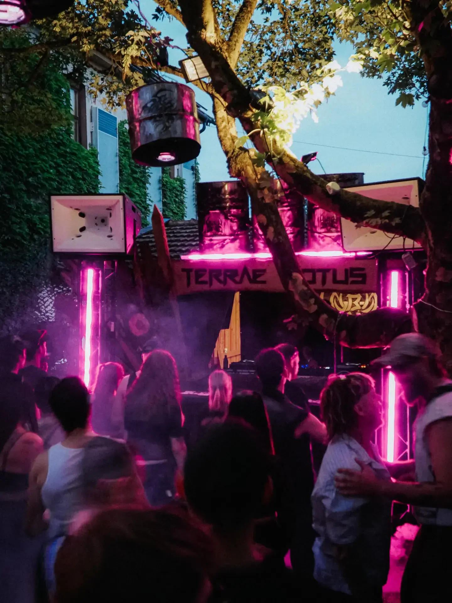 dieselbe bühne in magenta-neon bei einbruch der dunkelheit — tanzende crowd, hängende fass-lampen